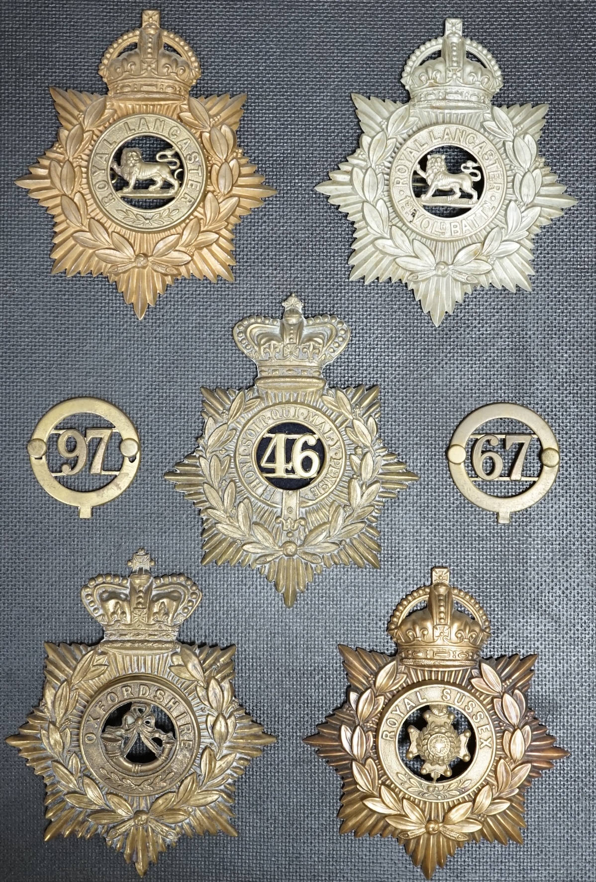 Five military helmet plates and two other centre badges mounted on a board, including; the Royal Lancashire Regiment, the Royal Lancashire 1st Volunteer Battalion, the Oxfordshire Regiment, the Royal Sussex Regiment, etc